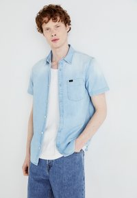 Chemise en denim bleu clair à manches courtes avec fermeture à bouton et poche poitrine, portée sur un débardeur blanc, associée à un jean bleu.