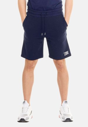 Man draagt navyblauwe shorts met trekkoord en het "LEONE"-logo op het linkerbeen en witte sportschoenen, staat met handen in de zakken.