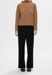 Pull en maille côtelée marron avec col montant, associé à un pantalon large noir et des chaussures noires sans lacets. Design simple et moderne.