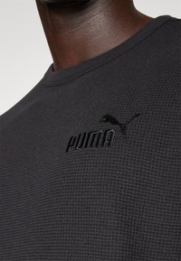 Schwarzes, strukturiertes Langarmshirt mit Rundhalsausschnitt, das eine stickerei mit "PUMA" und Logo in passender Farbe im Brustbereich zeigt.