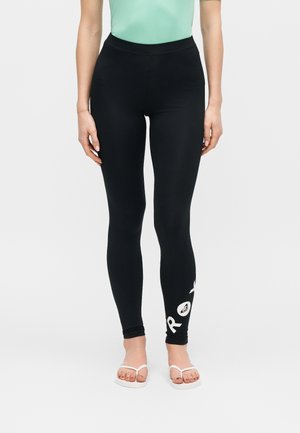 Naine kannab musti retuuse, millel on alumisel paremal jalal valge logo, ja valgeid sandaale, seisab sirgelt ühevärvilise taustaga ees.