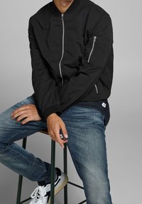 Svart bomberjacka med dragkedja framtill, ribbade muddar och krage samt dragkedjeförsedda sidofickor, bärs med blå jeans och vita sneakers.