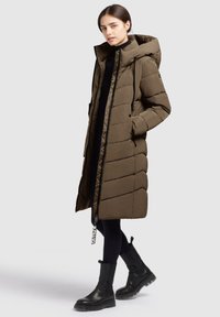 Naine, kes kannab pikka oliivroheline patjadega mantlit kapuutsiga, musti retuuse ja musti paksu talvesaapaid, seismas ühtlase tausta ees.