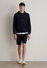 Marineblå hettegenser med frontlomme, sammen med svarte shorts og hvite sneakers. Modellen står mot en ensfarget bakgrunn.