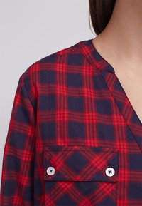 Rood en marineblauw geruite shirt met een v-hals ontwerp, borstzak en knoopsluitingen. De stof lijkt zacht met een gestructureerde afwerking.