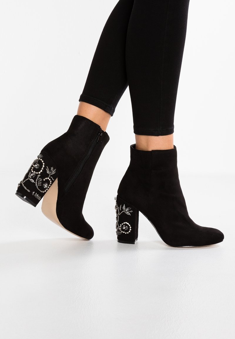Bottes chevilles en daim noir avec fermeture éclair, ornées de décorations florales sur un talon bloc. Texture lisse et design à bout pointu.