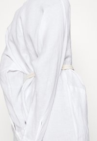 Il profilo laterale di una persona che indossa un lungo e fluente indumento bianco, stretto in vita con una corda di colore chiaro. Sfondo minimale.