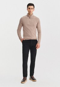 Beżowy sweter z prążkowanej tkaniny, z kołnierzem zapinanym na pół zamek, połączony z czarnymi spodniami i ciemnymi butami, charakteryzuje się dopasowanym krojem i teksturowanym materiałem.