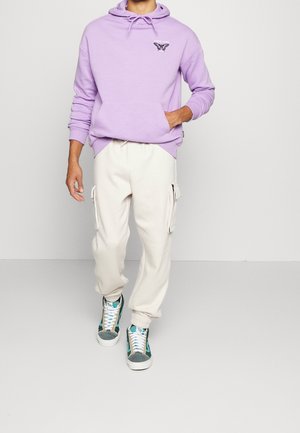 Felpa viola chiaro con tasca frontale e dettagli grafici; abbinata a pantaloni cargo crema con tasche con zip. Scarpe: design in teal e grigio.