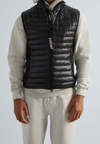 Gilet noir matelassé sans manches avec un quilting horizontal, fermeture éclair à l'avant et un détail de badge près de l'épaule, porté sur un sweatshirt léger gris.