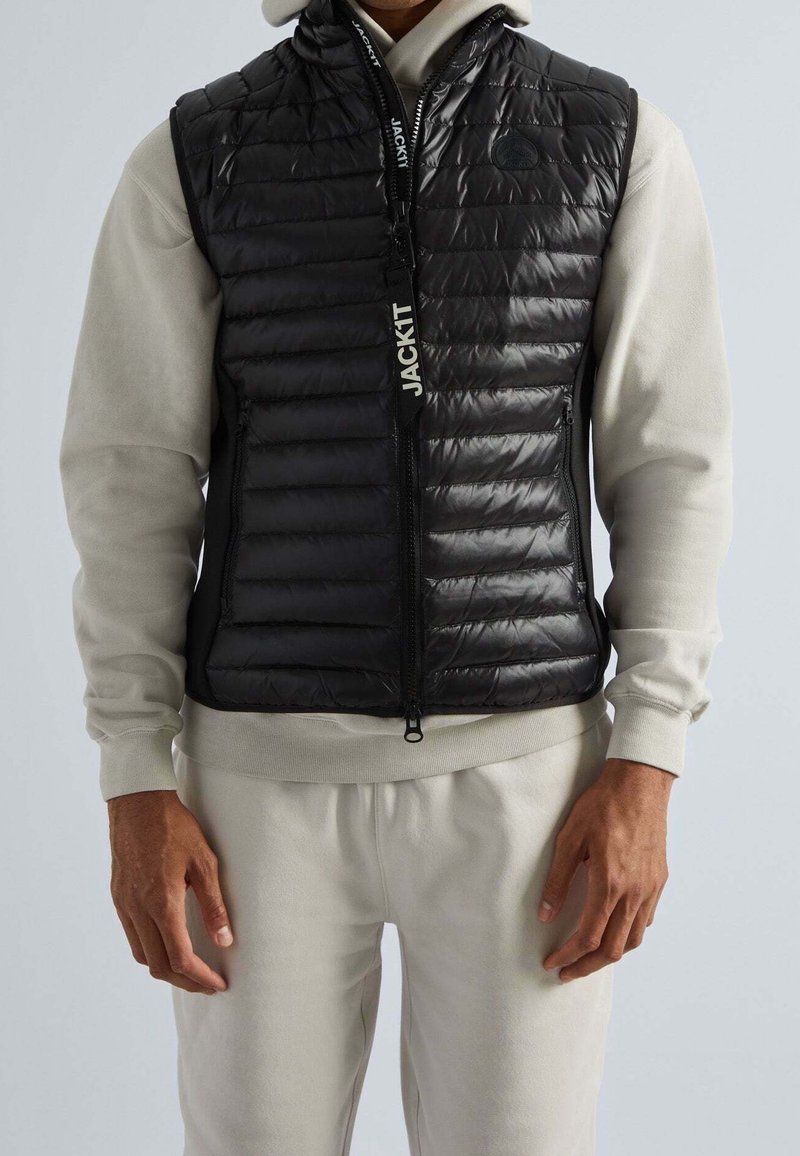 Gilet noir matelassé sans manches avec un quilting horizontal, fermeture éclair à l'avant et un détail de badge près de l'épaule, porté sur un sweatshirt léger gris.