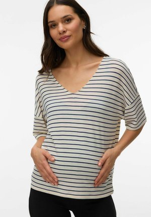 Kvinde iført en hvid og marineblå stribet V-hals skjorte, der blidt holder sin gravide mave med begge hænder, stående foran en hvid baggrund.