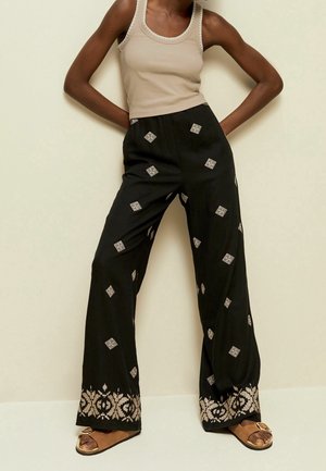 Femme portant un pantalon noir à jambes larges avec des motifs brodés beige, un haut sans manches beige et des sandales marron avec des boucles.