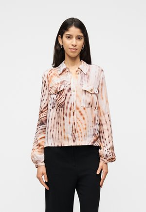 Donna che indossa una camicetta a maniche lunghe con bottoni, decorata con un motivo astratto marrone e beige, e pantaloni neri, in piedi contro uno sfondo semplice.