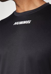 Nahaufnahme einer Person, die ein schwarzes Shirt mit rundem Ausschnitt und weißem "HUMMEL"-Logo trägt, welches zentriert unter dem Ausschnitt platziert ist.