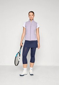 Ανοιχτό μωβ αμάνικο γιλέκο πάνω από ένα λευκό πουκάμισο, συνδυασμένο με μια ναυτική φούστα και αντίστοιχες κολάν. Κρατάει μια ρακέτα τένις. Λευκά αθλητικά παπούτσια.