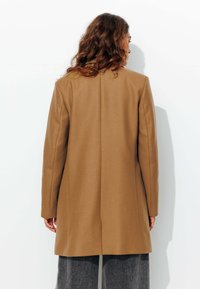 Manteau marron au design ajusté, avec une coupe droite, des manches longues et un tissu texturé. Minimaliste, sans motifs ni accents visibles.