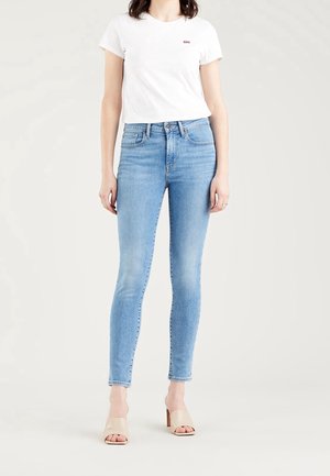 Femme portant un t-shirt blanc à manches courtes, un jean skinny bleu clair et des sandales à talons beiges, debout devant un fond clair uni.