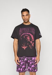 Sort t-shirt med stort pink grafiktryk med et kranium og tekst, parret med lilla mønstrede shorts med kranier.