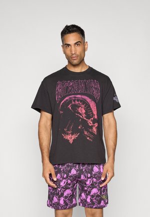 Sort t-shirt med stort pink grafiktryk med et kranium og tekst, parret med lilla mønstrede shorts med kranier.