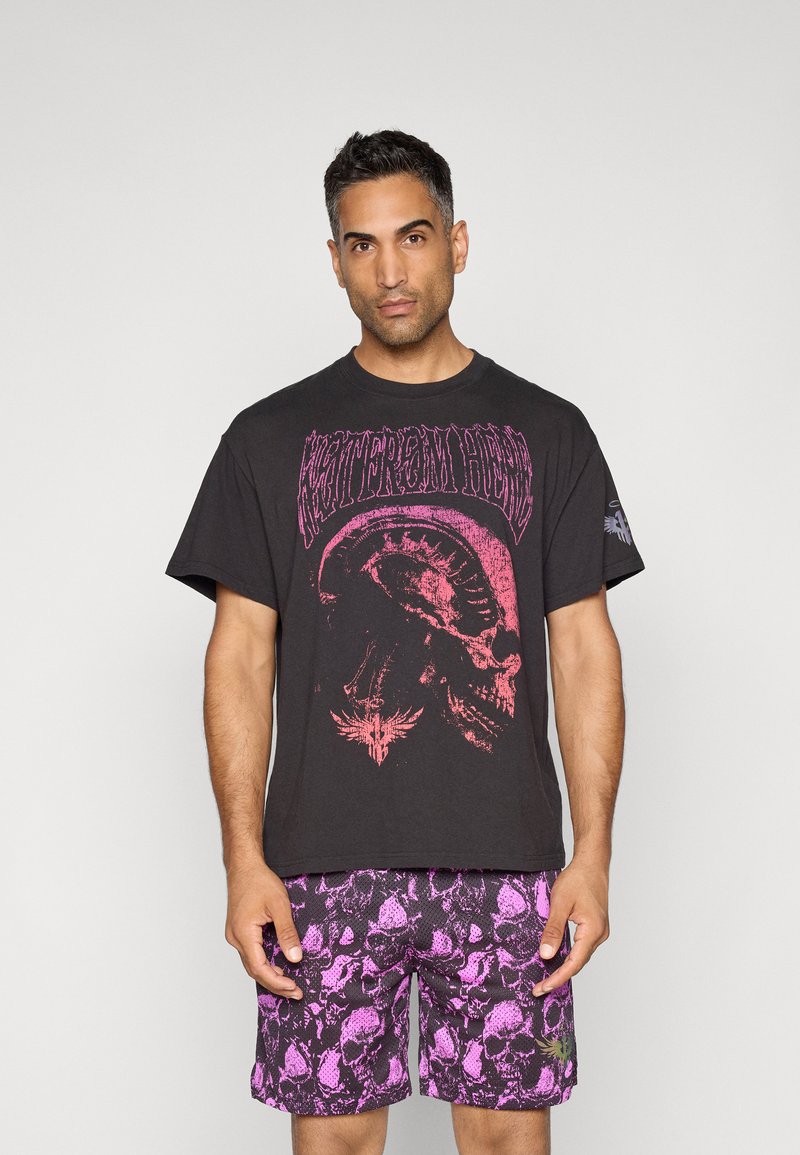 Sort t-shirt med stort pink grafiktryk med et kranium og tekst, parret med lilla mønstrede shorts med kranier.