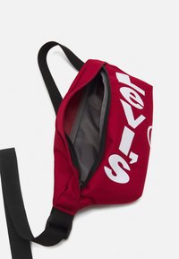 Sac en tissu rouge avec une sangle noire, grand logo blanc "Levi's", fermeture éclair, et doublure intérieure grise. Forme globale compacte et angulaire.
