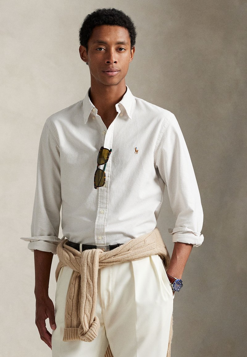 Camisa branca às riscas com botões, suéter bege de malha entrançada amarrado à cintura, calças claras, óculos de sol pendurados na camisa, relógio no pulso.