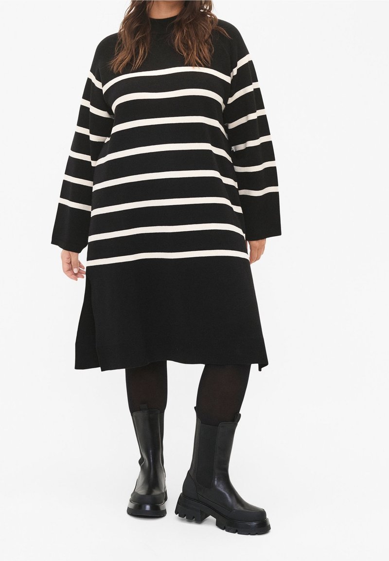 Schwarz-weiß gestreiftes, übergroßes Kleid mit langen Ärmeln und seitlichen Schlitzen, kombiniert mit kniehohen, schwarzen Boots mit dickem Sohlen.