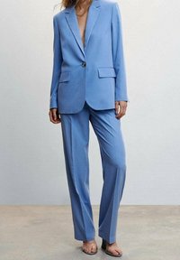 Costume bleu clair avec un blazer sur mesure à revers et deux poches avant, accompagné d'un pantalon droit, les deux en tissu lisse.