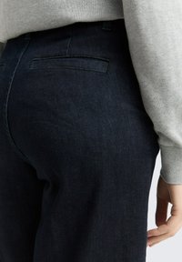 Jean en denim bleu foncé à texture lisse, présentant un empiècement en forme de selle et deux poches arrière. Le vêtement est assorti à un pull gris clair.
