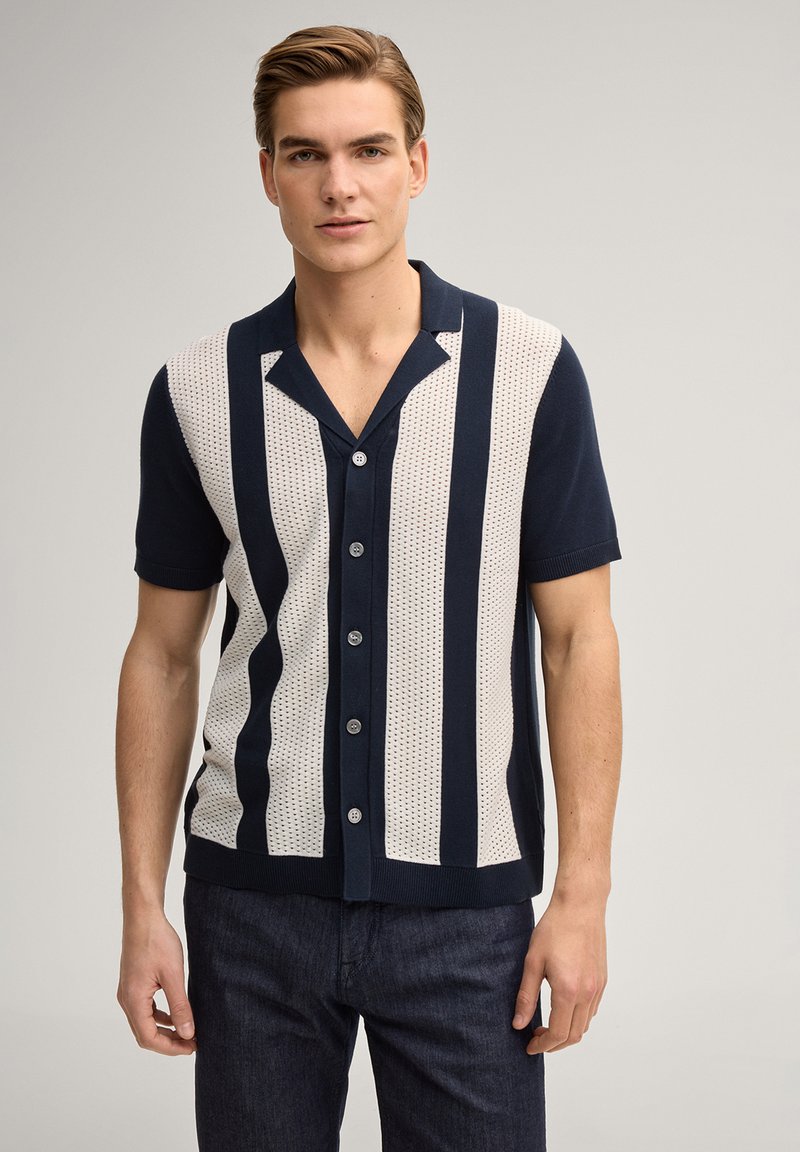 Young man wearing a navy and white short-sleeve button-up shirt with vertical stripes, standing against a plain background.