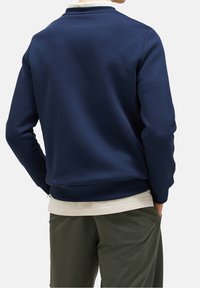 Sudadera azul marino hecha de una mezcla de algodón, con un cuello redondo, puños acanalados y un corte relajado, llevada sobre una camisa crema.