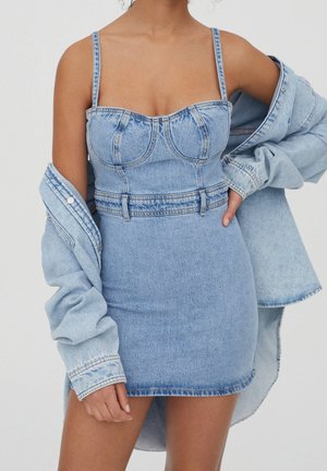 Kobieta ubrana w jasno niebieską dżinsową sukienkę typu bustier z cienkimi ramiączkami i dżinsową kurtkę opadającą z ramion na jednolitym tle.