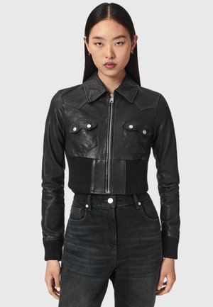 Frau trägt eine schwarze, kurze Lederjacke mit Frontreißverschluss und knöpfbaren Brusttaschen, kombiniert mit hoch taillierten schwarzen Jeans.