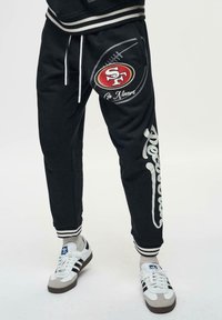 Zwarte sweats met een trekkoord in de taille, met het "SF" logo en de tekst "Pro Niners", witte accenten en gestreepte cuffs. Textuurstof.