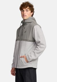 Gray fleece jacket with a hood, contrasting collar, and patterned chest stripe. Features front pockets and a zipper closure. Textured fabric.