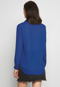 Blå långärmad blus med krage, slät textur, rundad nederkant och knäppta ärmslut. Bärs tillsammans med en svart denimkjol med fransade kanter.