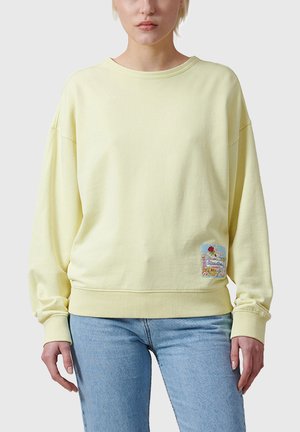 Persona indossa una felpa giallo chiaro con collo a giro e una toppa colorata vicino all'orlo inferiore, abbinata a jeans azzurri.