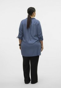 Tunique bleu à manches longues avec des manches retroussables, en tissu lisse et coupe décontractée, associée à un pantalon large noir. Vue arrière simple.