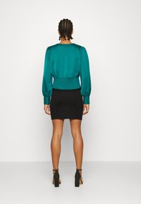 Blusa di satin turchese con maniche a sbuffo e vita arricciata, abbinata a una minigonna nera. La modella indossa scarpe con tacco a blocco nere.