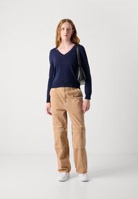 Maglione blu a V abbinato a pantaloni cargo beige con tasche a toppa, completato da sneakers bianche e una piccola borsa nera.