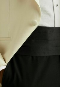 Kermainen tuxedo-takki pyöreällä napilla, yhdistetty mustan vyötärönauhan ja housujen kanssa. Vaatekokoonpanossa näkyy kangaspintojen tekstuurity yksityiskohtia.