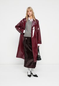 Burgunderfarvet vinyl trenchcoat over en grå kabelstrikket sweater, parret med en mørk nederdel med paljetter, hvide sokker og sorte hæle, mens der holdes en lille taske.