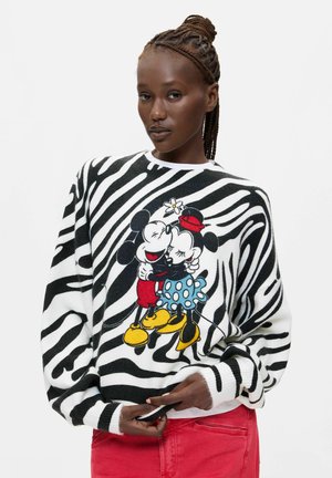 Kobieta z zaplecionymi włosami nosi sweter w paski zebry z Mickey i Minnie Mouse obejmującymi się, do tego czerwone spodnie.