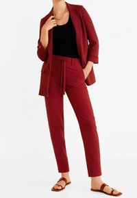 Blazer bourgogne avec manches retroussées, associé à un débardeur noir et un pantalon ajusté bourgogne assorti. Des sandales plates marron complètent la tenue.