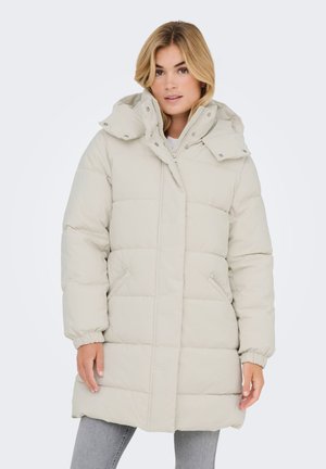 Woman wearing a light beige quilted puffer coat with a high collar and hood, paired with gray jeans on a plain white background.