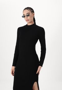 Abito nero a costine con scollo alto e maniche lunghe. Presenta uno spacco laterale per un tocco in più. Indossato con occhiali da sole sottili.