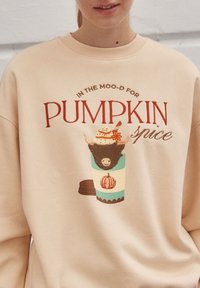 Crèmekleurige sweatshirt met een grafisch ontwerp van een pompoenkruidendrank, decoratieve elementen en speelse tekst in rode en bruine tinten.