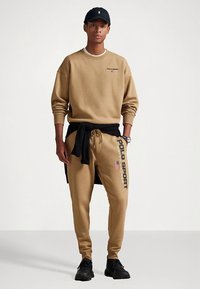 Μπεζ φούτερ με κεντημένο λογότυπο "POLO SPORT", συνδυασμένο με ταιριαστό παντελόνι φόρμας με τυπωμένο λογότυπο και αμερικανική σημαία. Μαύρο καπέλο και αθλητικά παπούτσια.