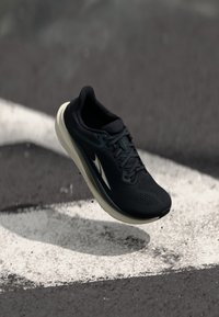Juodas bėgimo batelis su tinklelio viršumi, „Flyknit“ tekstūra ir kontrastuojančia smėlio spalvos padu. Yra apvalus pirštas ir tradicinis raištelių užsegimas.